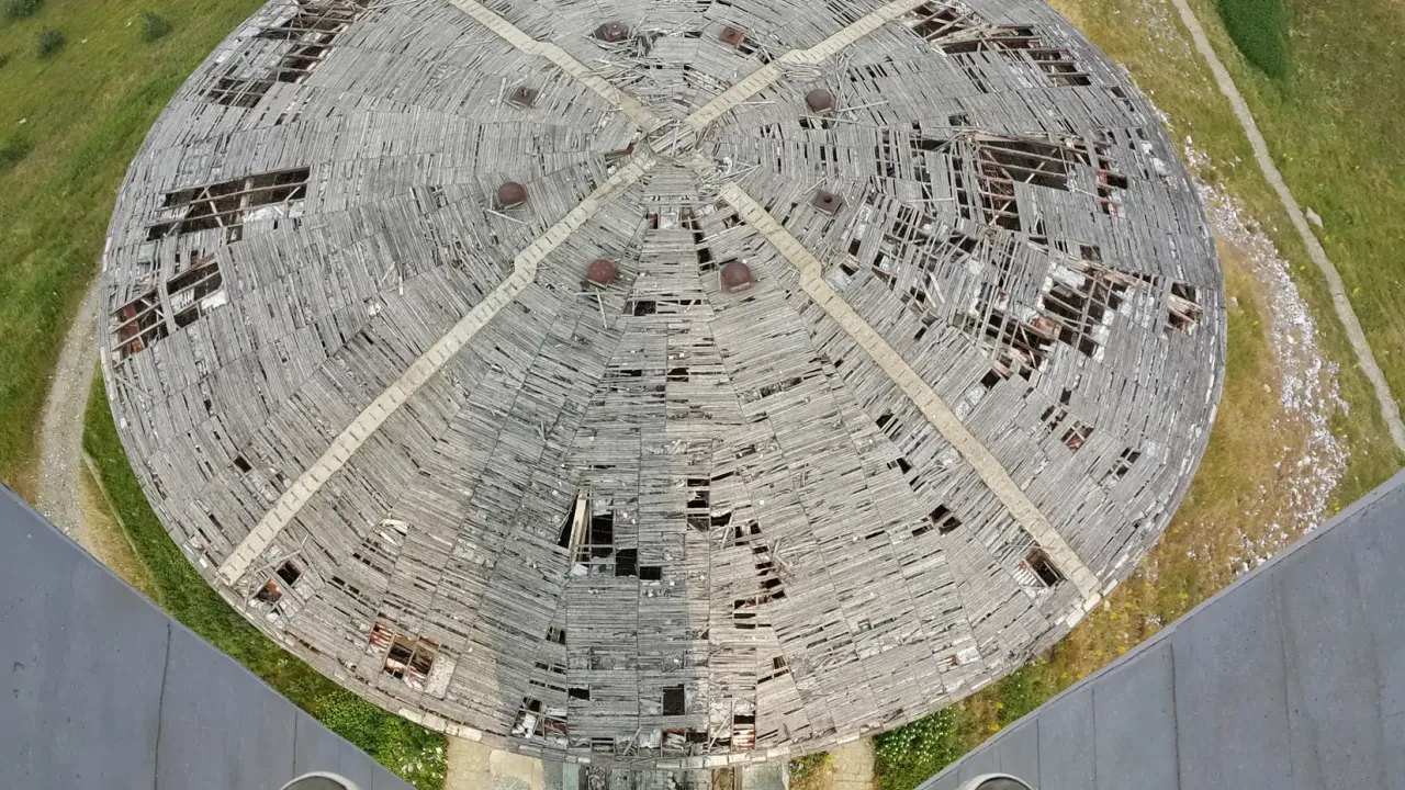 Et svimlende kig ned fra tårnet mod Buzludzha-monumentets runde, beskadigede tag; fotografens fødder ses i forgrunden.
