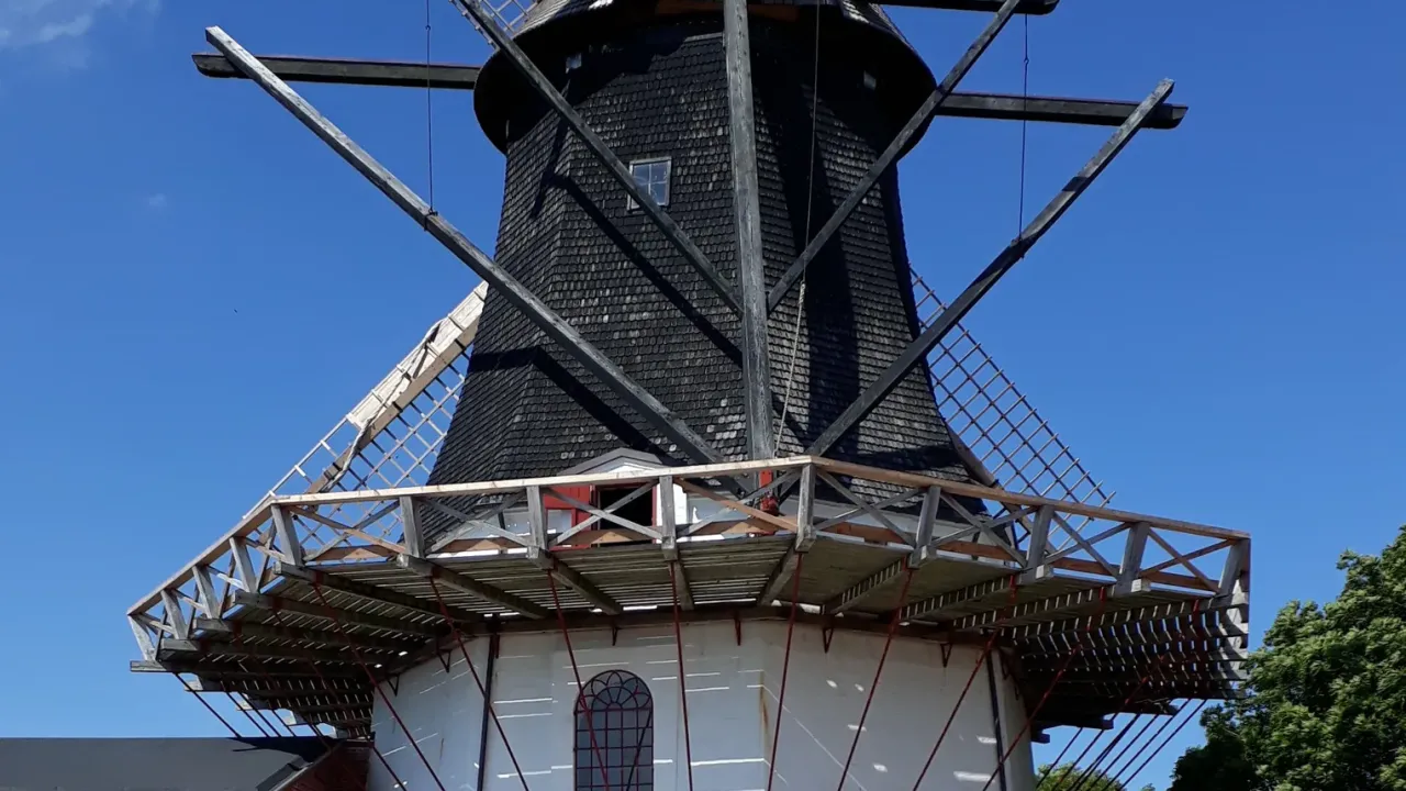 Højer Mølle med mørk træbeklædning, hvid underbygning og vinger mod blå himmel.