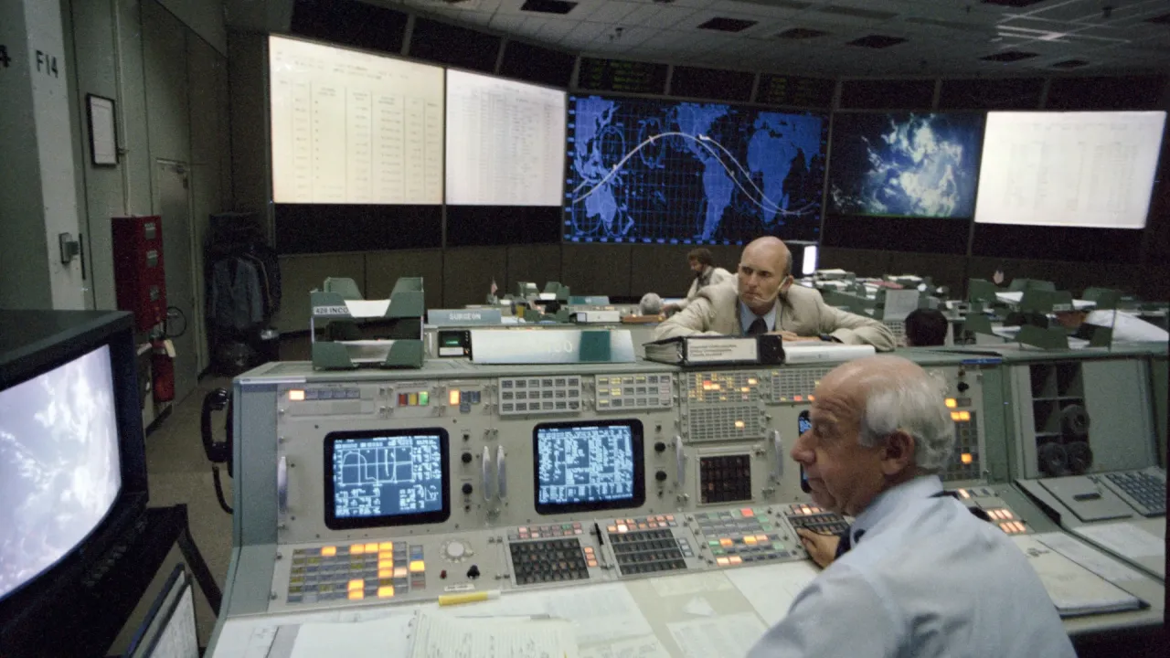 C. Gordon Fullerton læner sig over en konsol, mens Edward Fendell sidder foran skærme i NASAs kontrolrum i Houston.