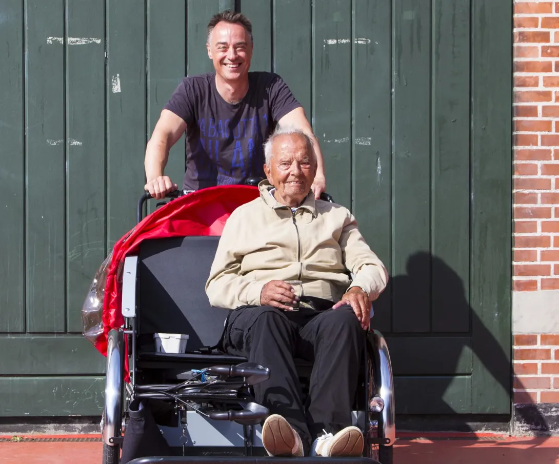 Ole Kassow står bag en rickshaw med Thorkild Thim på passagersædet foran en grøn port.