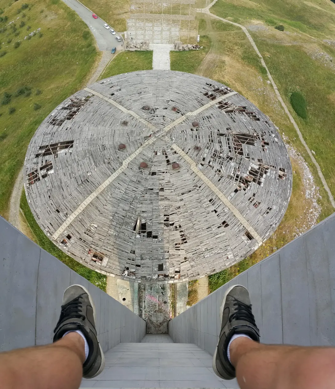 Et svimlende kig ned fra tårnet mod Buzludzha-monumentets runde, beskadigede tag; fotografens fødder ses i forgrunden.
