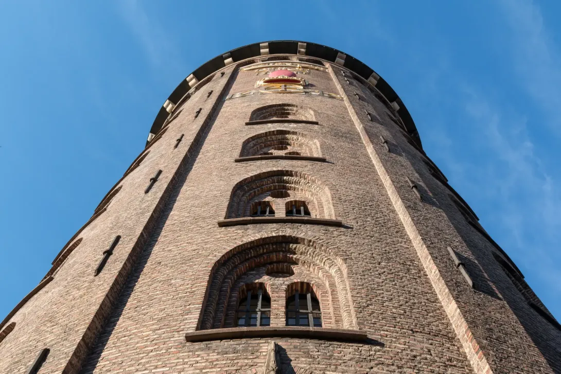 Rundetårns runde murstensfacade og gyldne rebus set stejlt nedefra mod en blå himmel.