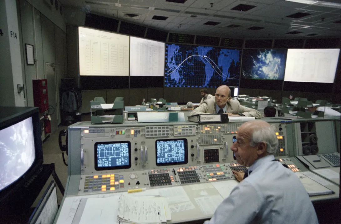 C. Gordon Fullerton læner sig over en konsol, mens Edward Fendell sidder foran skærme i NASAs kontrolrum i Houston.
