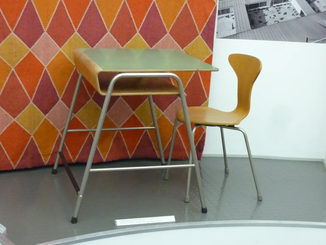 Arne Jacobsens skolebord og en gul stol fra Munkegårdsskolen, udstillet på Designmuseum Danmark foran et farvet tekstil.