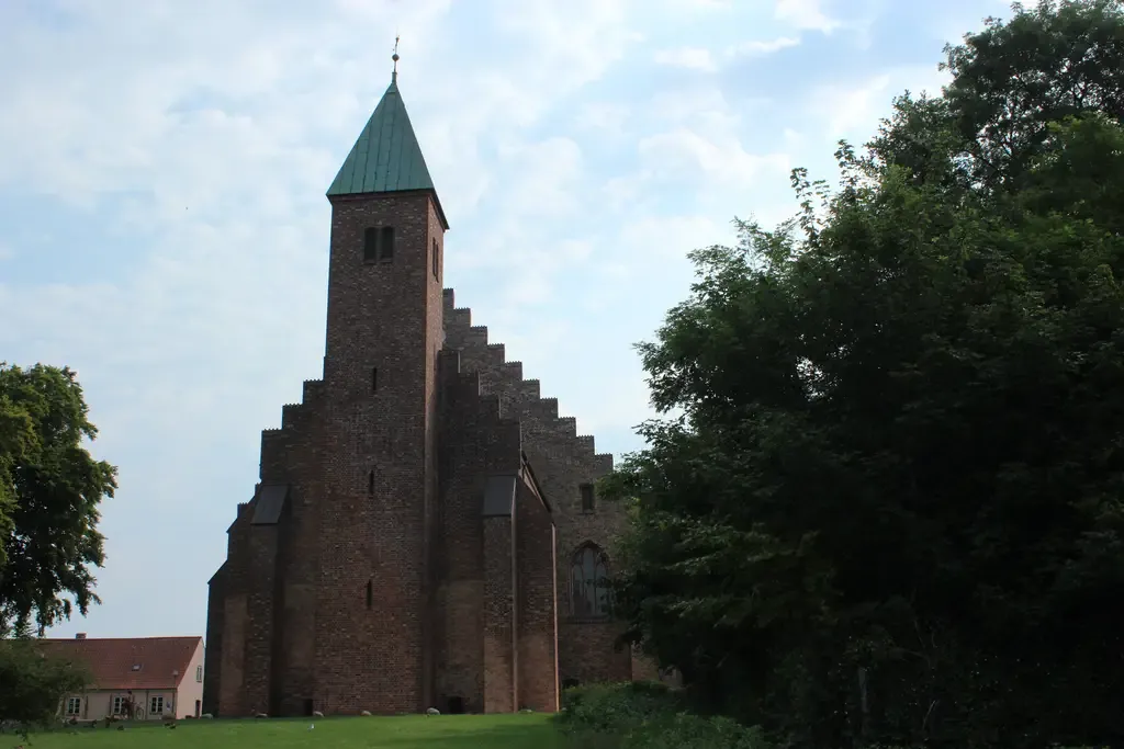 Maribo Domkirke med røde teglstensgavle og et slankt tårn set fra søsiden mellem grønne træer.